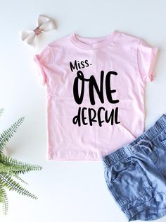 a pink shirt that says, miss onederful next to some denim shorts and a plant