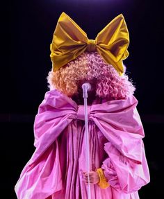 a drag queen dressed in pink and yellow with a big bow on her head, holding a microphone