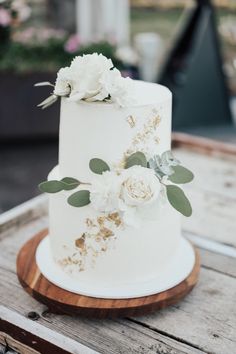 a wedding cake with white flowers and greenery on top sitting on a wooden table