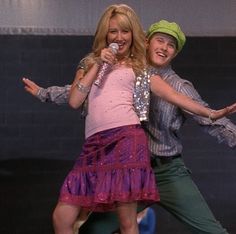 two people on stage one is holding a microphone and the other is wearing a skirt