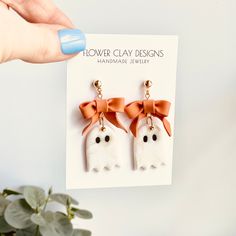 a pair of white and orange bow earrings with black eyes on them, hanging from a card that says flower clay designs