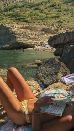 a woman laying on top of a towel next to the ocean with an orange book in her hand