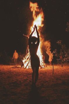 a woman standing in front of a fire with her arms up and hands raised above her head