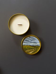 two candles sitting next to each other on top of a gray surface with a painting in the middle