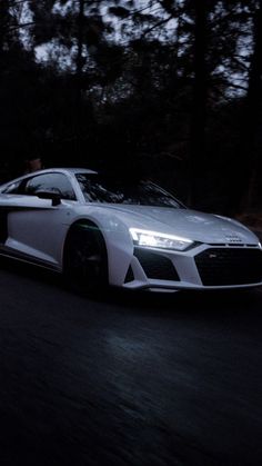 a white sports car driving down the road at night with its headlights on and trees in the background