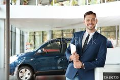 a man in a suit and tie standing next to a blue car at a dealership