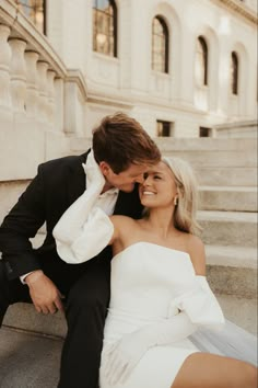 a man and woman are sitting on the steps in front of a building, smiling at each other