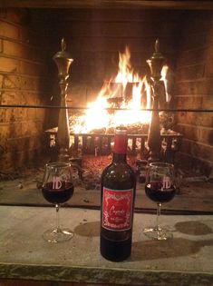 two wine glasses sitting in front of an open fire place with a bottle on the table