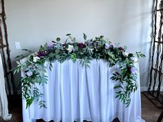 an arrangement of flowers and greenery on top of a white table cloth draped over it