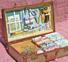 an open suitcase filled with sewing supplies on a pink striped cloth covered surface, next to scissors and thread