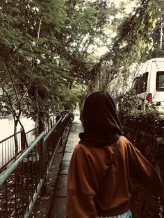 a woman in a brown hoodie walking down a sidewalk next to a fence and trees