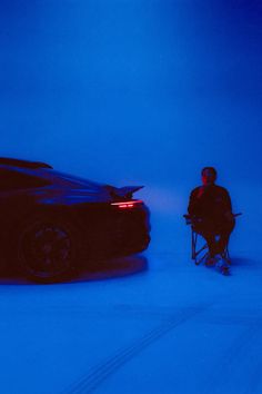 a man sitting in a chair next to a sports car on the snow covered ground