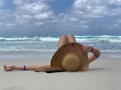 a woman laying on top of a sandy beach next to the ocean wearing a straw hat