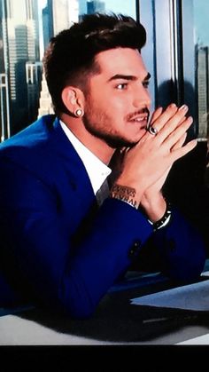 a man in a blue suit sitting at a table with his hands clasped to his chest