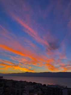 the sky is very colorful at sunset over some buildings by the water and mountains in the distance