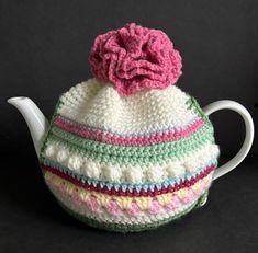 a crocheted tea pot cover with a pink flower on the top and green, blue, yellow, white, and pink stripes