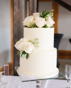 a wedding cake with white flowers and greenery sits on a glass stand at the head table