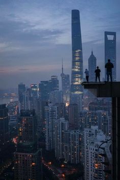 two people standing on top of a tall building looking at the city lights and skyscrapers