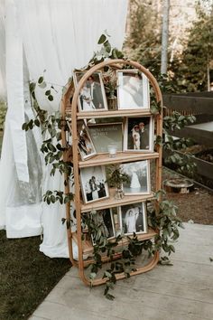 a wooden book shelf with pictures and greenery on it in front of a white curtain