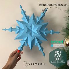 a hand holding a blue paper snowflake next to a christmas tree