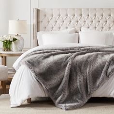 a bed with white linens and a gray fur blanket on it's headboard