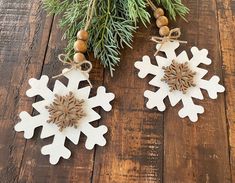 two wooden snowflakes hanging from a tree branch on a wood table with pine cones and twine