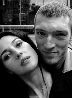 a black and white photo of a man and woman posing for the camera with buildings in the background