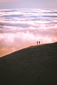 two people standing on top of a hill above the clouds