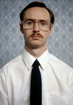 a man with glasses and a mustache wearing a white shirt, black tie and gold rimmed glasses