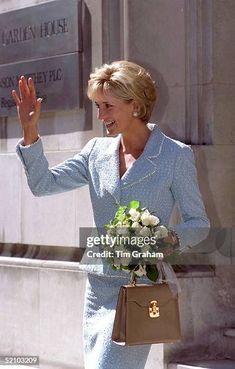 a woman in a blue dress waving and holding her hand out to the side while standing outside