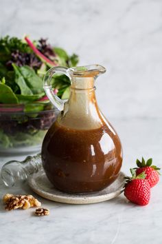 a glass bottle filled with brown liquid next to a bowl of greens and a strawberry