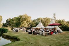 a group of people sitting at tables in the grass near a pond and tents with red umbrellas