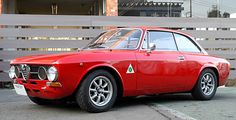 an old red sports car parked in front of a wooden fence on the side of a road