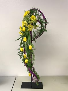 a vase with yellow flowers and greenery on a black stand in front of a white wall