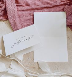 two white envelopes sitting on top of each other next to a pink blanket and flowers