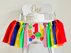 a white chair topped with a cake covered in rainbows and apples next to a one year old birthday cake