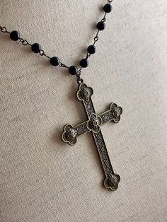 Step back in time with this intricate hand-assembled metal cross rosary-style necklace, featuring a long black beaded chain, and quatrefoil design bronze colored cross pendant. Perfect for tying together a Gothic Medieval, or perhaps Victorian Mourning styled look. This design is part of our handmade collection, making each piece unique. Sizing: One SizeChain Length - 30 inches / 76 CentimetersPendant Length - 3 inches / 7.6 CentimetersTotal Length Laid Flat - 18 inches / 45.5 Centimeters Care I Bronze Cross Bohemian Necklace, Bronze Bohemian Cross Necklace, Bohemian Bronze Cross Necklace, Medieval Black Metal Necklace, Medieval Style Black Metal Necklace, Handmade Black Crucifix Necklace, Black Spiritual Jewelry With Antique Finish, Gothic Black Brass Necklace, Black Gothic Brass Necklace