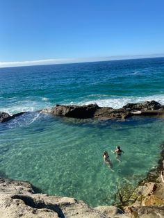 two people are swimming in the clear blue water at the edge of an ocean cliff