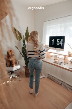 a woman standing in front of a desk with a computer on it and a dog sitting next to her