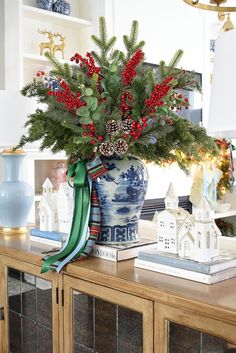 a vase filled with red berries and greenery on top of a wooden table next to a christmas tree