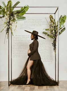 a woman in a long black dress and hat is sitting on a bench with plants behind her