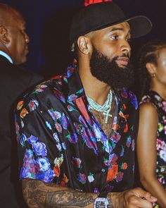 a man with a beard wearing a floral shirt and black hat sitting in front of other people