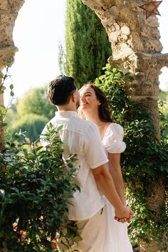 a man and woman standing next to each other in front of an arch with greenery