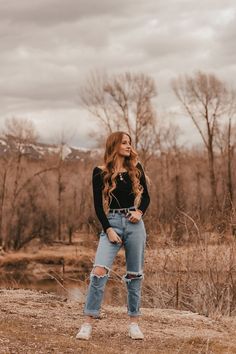 a woman standing on top of a dirt field wearing jeans and a black shirt with ripped knees