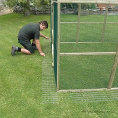 a man kneeling down next to a tall green chicken coop on top of a lush green field