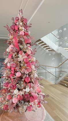 a pink christmas tree with lots of ornaments on it in front of a stair case