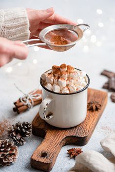 someone pouring hot chocolate into a mug with marshmallows and cinnamon on top