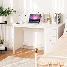 a laptop computer sitting on top of a white desk next to a bed and potted plant