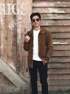 a man standing in front of a wooden building wearing sunglasses and a brown suede jacket