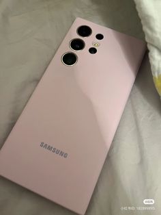 a pink samsung phone laying on top of a white bed next to a yellow pillow